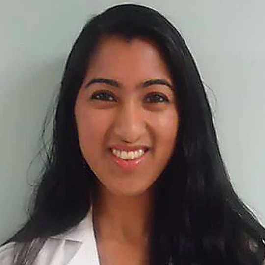 A person with long black hair smiling, wearing a white top, against a plain background.