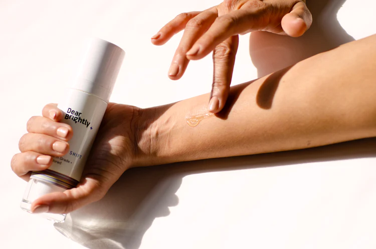 A person applies a cream from a "Dear Brightly" bottle onto their arm using a finger, set against a plain white background.